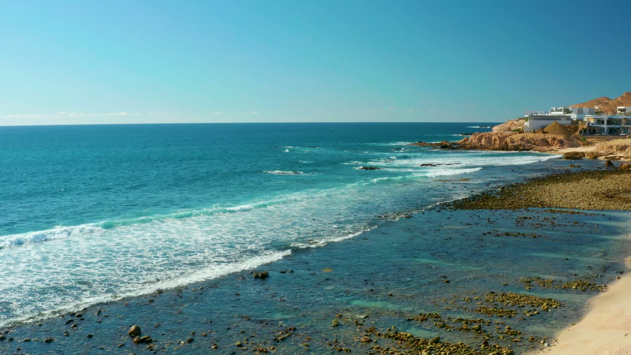 White surf rolls across shallow rocks near a sandy beach and cliff buildings.