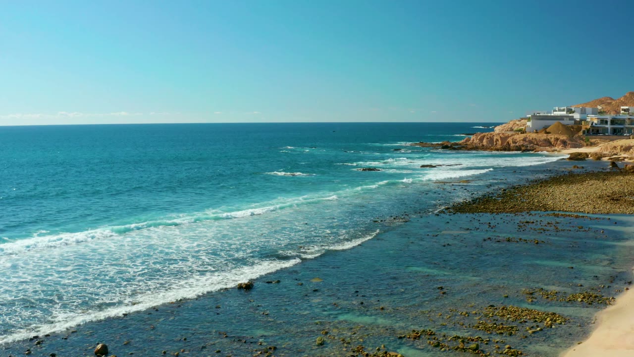 At 0:02 in El Tule Los Cabos, the surf line curves diagonally across clear blue water toward the rocky point and cliff buildings, with no visible people.