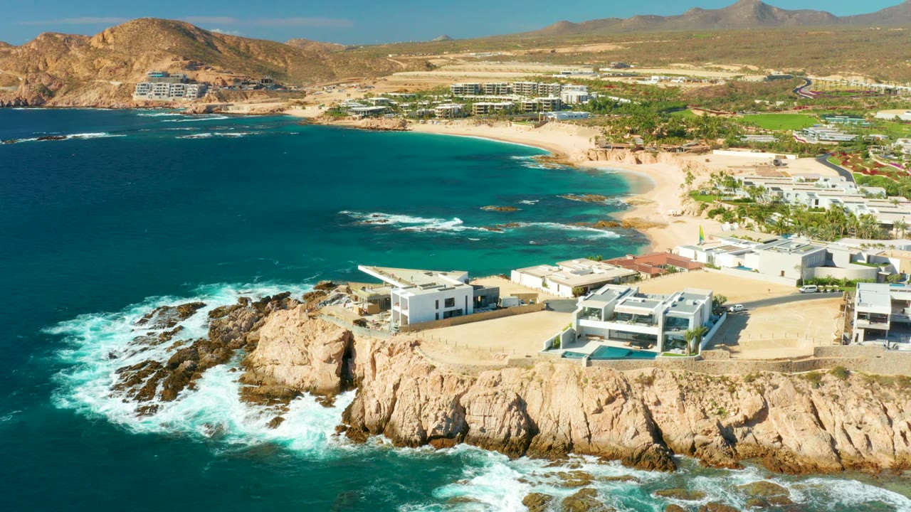 Aerial frame over Chileno Beach in Los Cabos showing turquoise water on the left, tan cliffs below, modern white villas near the edge, sandy beach beyond, and no visible people.