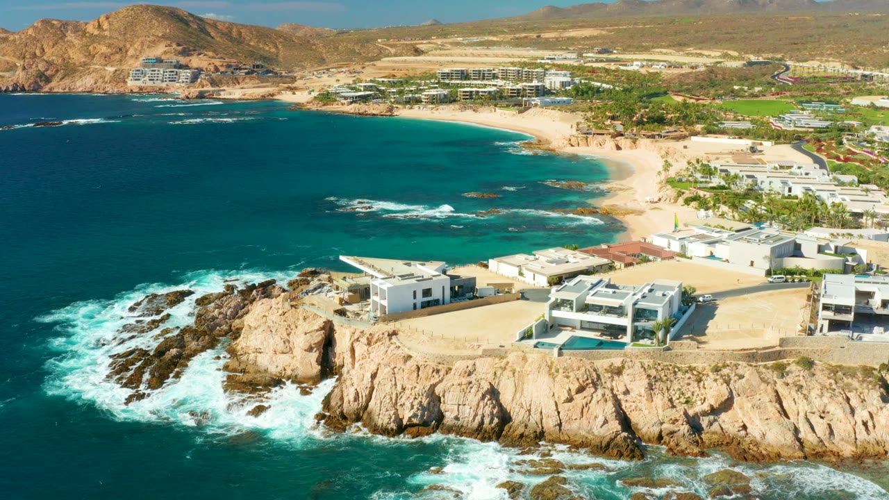 Aerial coastline with surf breaking on rocks beside white modern buildings and a sandy beach.