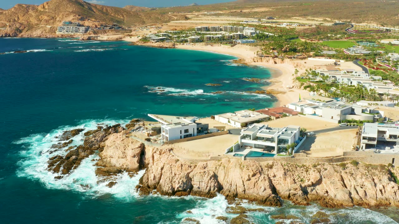 Wide aerial frame at Chileno Beach in Los Cabos showing the blue-green bay filling the left side, surf on the rocks below, villas along the cliff, beach in the midground, and no visible people.