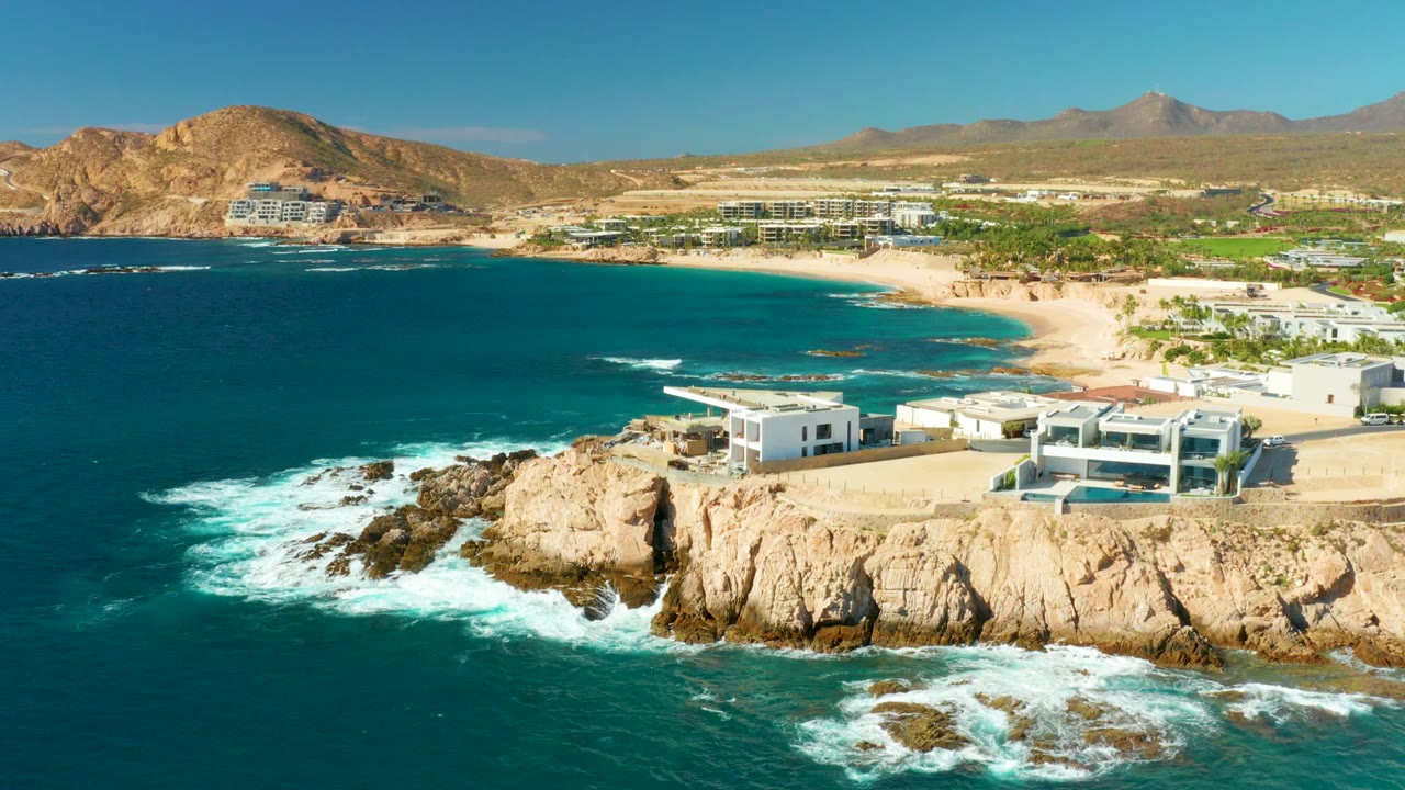 Opening frame over Chileno Beach Los Cabos shows turquoise water and white surf beside tan rocky cliffs, beachfront buildings, sandy beach, desert hills, and no visible people.