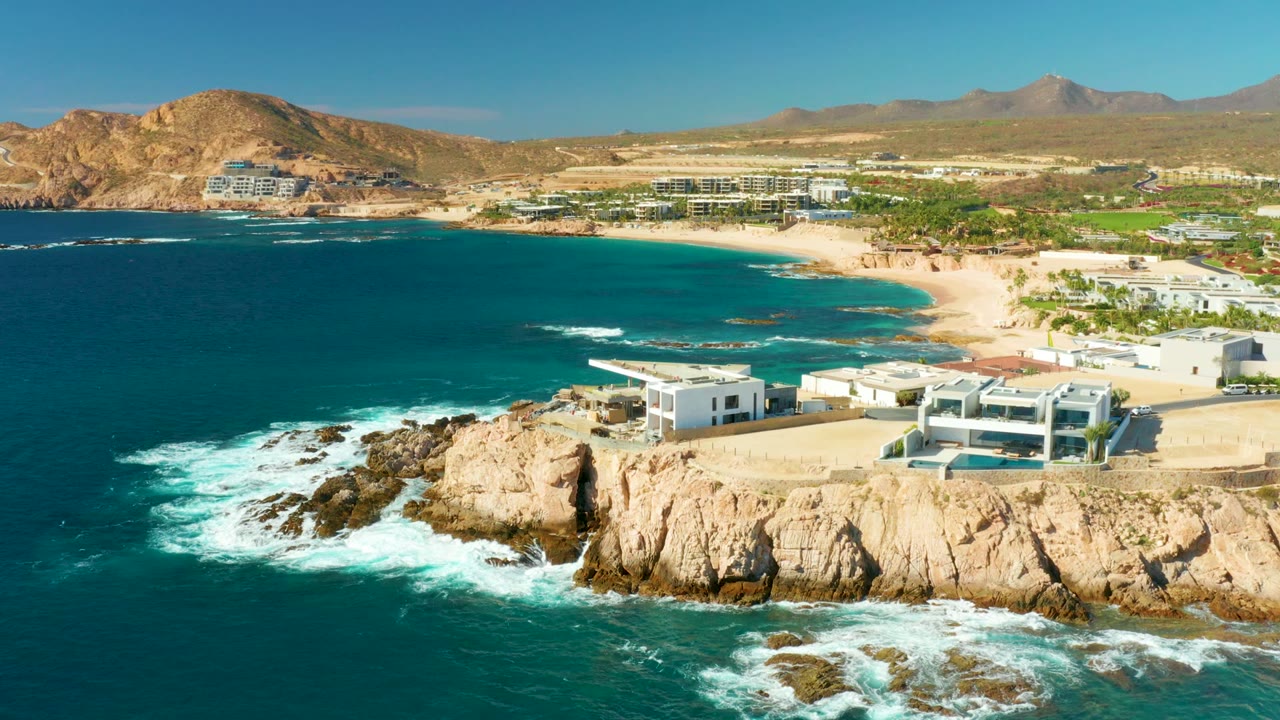 Wide drone view of waves breaking on rocks below beachfront buildings and a sandy cove.
