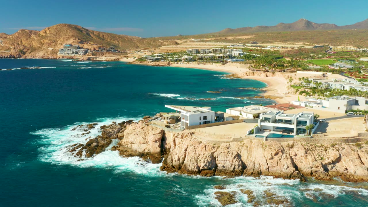 At 0:02, the aerial view shows the rocky point and white surf slightly farther across frame, with Chileno Beach buildings, palms, sand, arid hills, and no visible people.