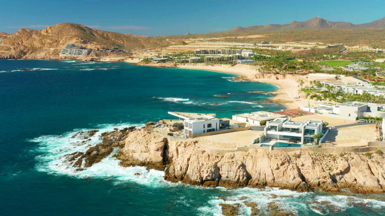 Aerial coastal scene with cliffs in foreground, blue water, beach, buildings, and hills.