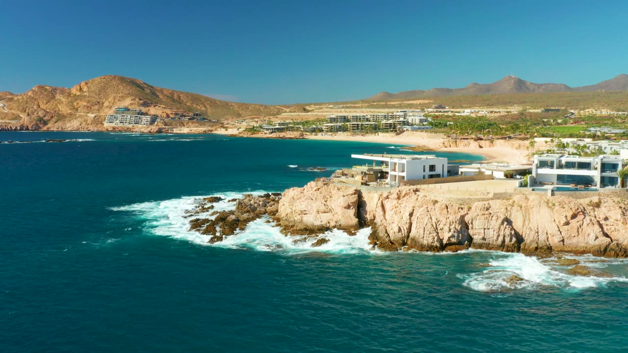 Aerial view over Chileno Beach in Los Cabos, with turquoise water and white surf hitting tan rocks below beachfront buildings, sandy beach, dry hills, and no visible people.