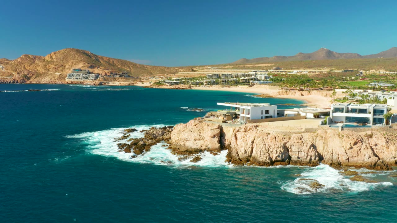 Waves breaking around rocks below white buildings and a sandy beach