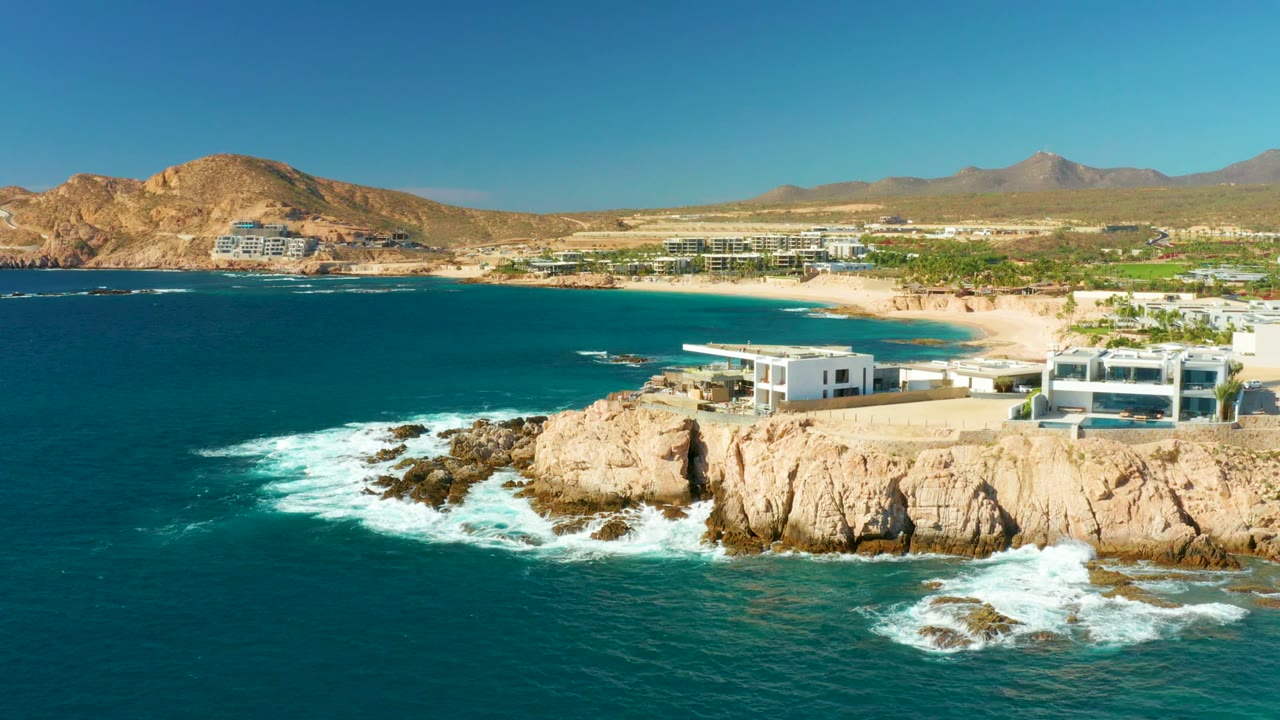 Chileno Beach remains in view as the surf shifts lower around the rocks, with a modern white building near the cliff edge, resort-area buildings inland, mountains, and no visible people.