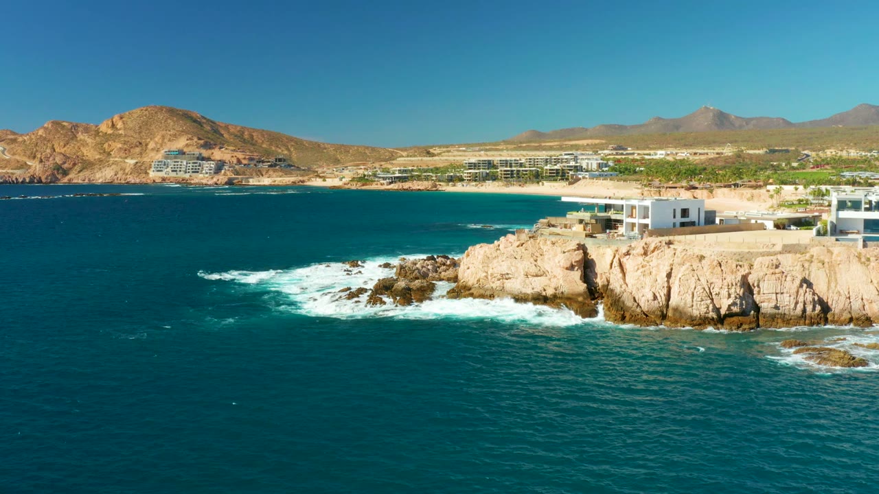 At Chileno Beach Los Cabos, turquoise water and white foam meet tan coastal rocks, with modern white cliffside buildings at right, dry hills and mountains beyond, and no visible people.