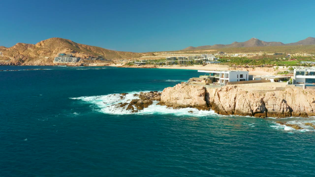 At 0:02, Chileno Beach Los Cabos appears from an elevated coastal angle as white surf curls around the rocks, the cliffside buildings sit near the right edge, and no people are visible.