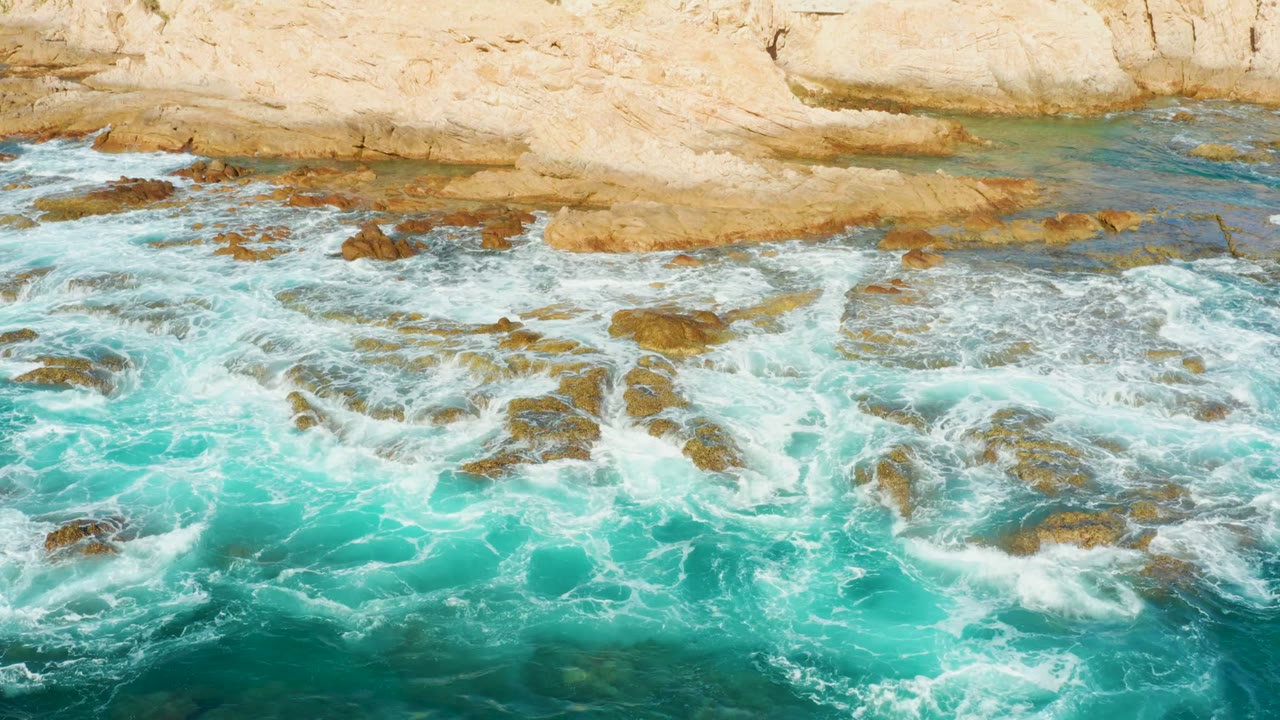 At Chileno Beach in Los Cabos, turquoise surf spreads white foam over exposed rocks beneath a tan cliff, with no visible people.