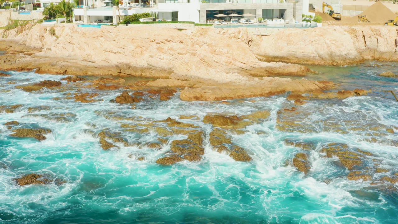 At Chileno Beach in Los Cabos, waves break around shoreline rocks while the cliff edge and parts of white modern buildings appear above, with no visible people.