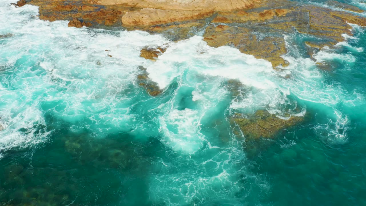 A dense patch of white foam churns over shallow brown rocks at Chileno Beach in Los Cabos as clear turquoise water surrounds it, with no visible people.