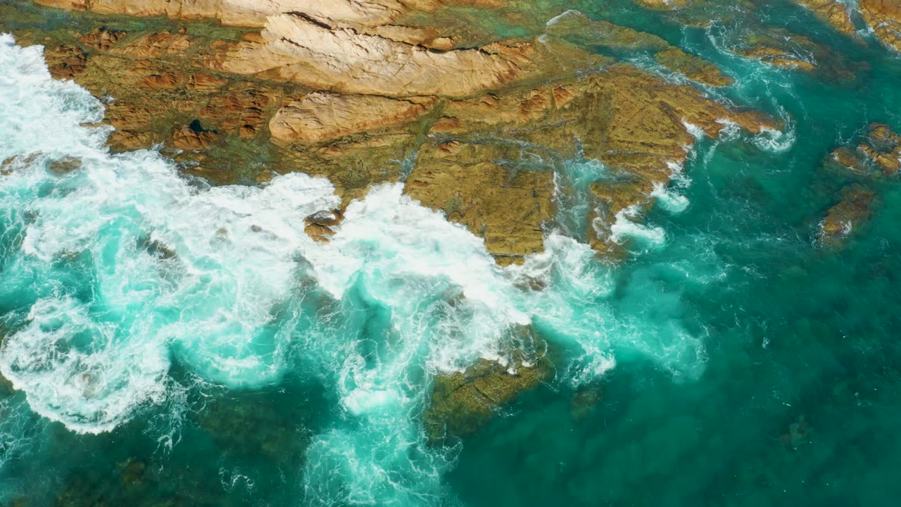 Overhead view of turquoise surf spreading across brown coastal rocks at Chileno Beach in Los Cabos, Mexico, with no visible people.