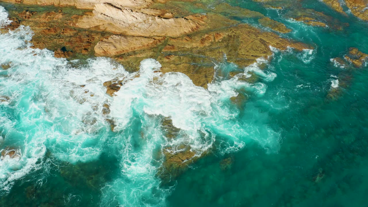 Top-down view of breaking surf spreading wider across flat tan and brown rocks at Chileno Beach in Los Cabos, Mexico, with no visible people.