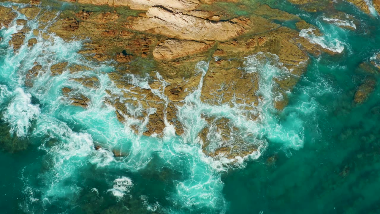 Overhead view at Chileno Beach in Los Cabos showing turquoise water and white foam breaking over tan rocky ledges, with no visible people.