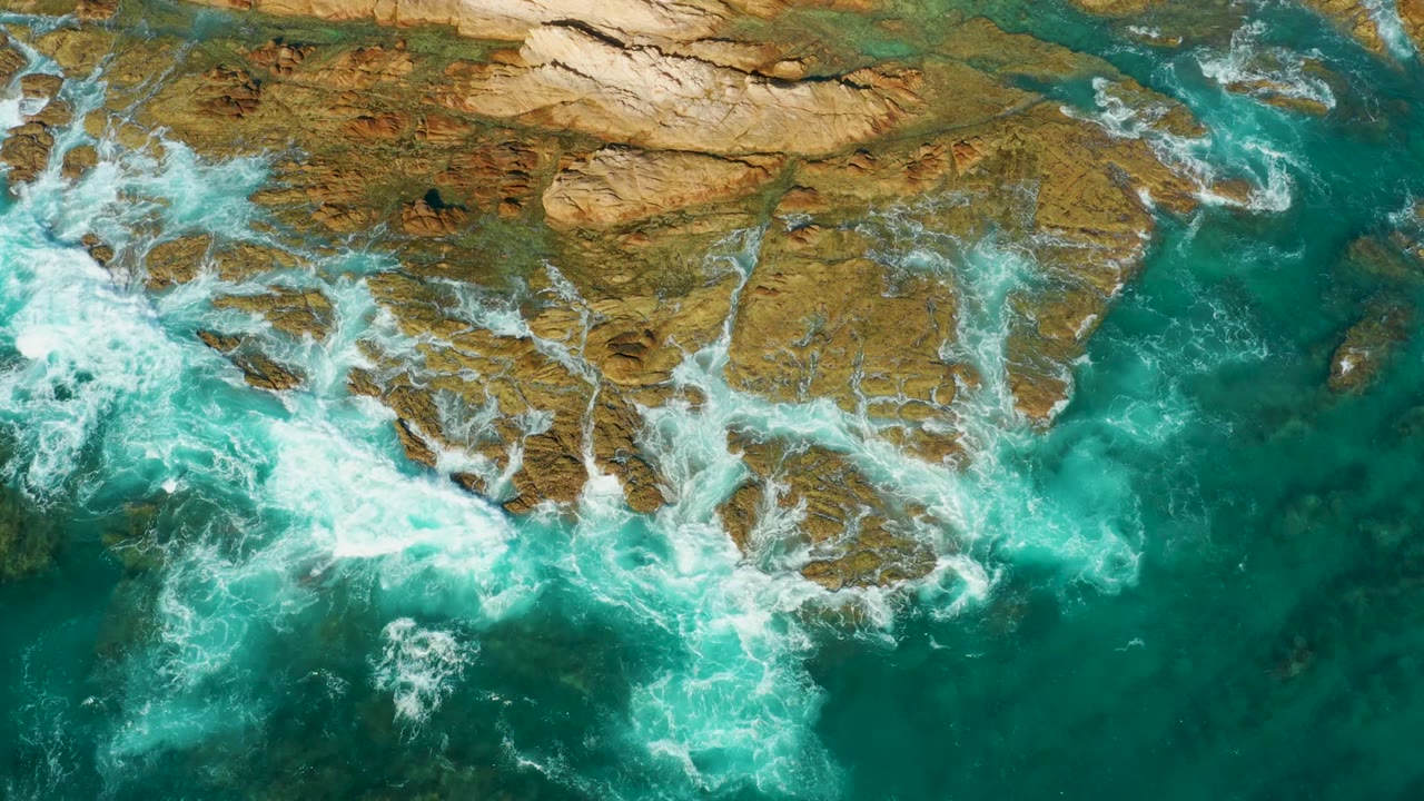 Overhead surf at Chileno Beach curls around uneven tan and brown coastal rocks as deeper turquoise water sits beside the ledges, with no visible people.
