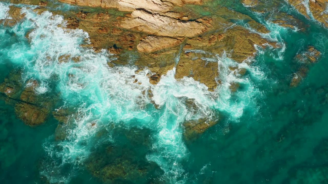 At Chileno Beach in Los Cabos, turquoise water pushes white foam over tan and brown rocks near the top of the frame, with no visible people.