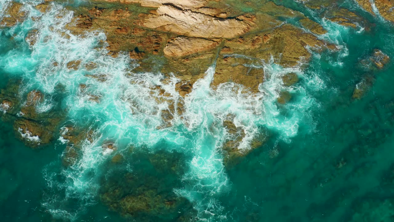 At Chileno Beach in Los Cabos, foamy water curls around exposed rock points and leaves a deeper turquoise channel on the right, with no visible people.