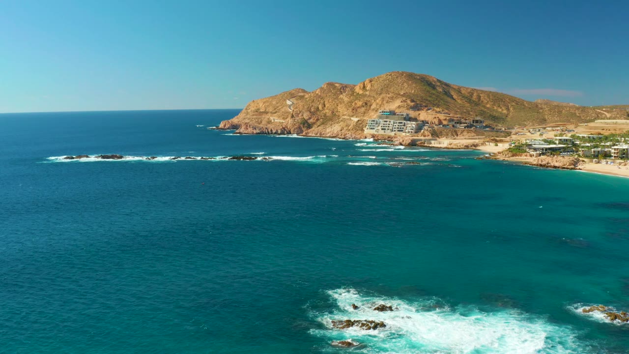Opening aerial frame over Chileno Beach Los Cabos showing turquoise ocean, white surf on rocky outcrops, a sandy beach edge, hillside buildings, tan hills, blue sky, and no visible people.