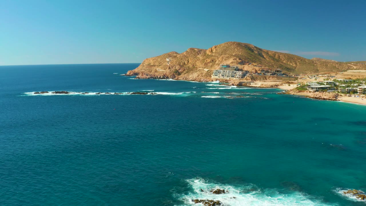 Wide drone view of turquoise water, rocky surf, beach, hillside, and coastal buildings.