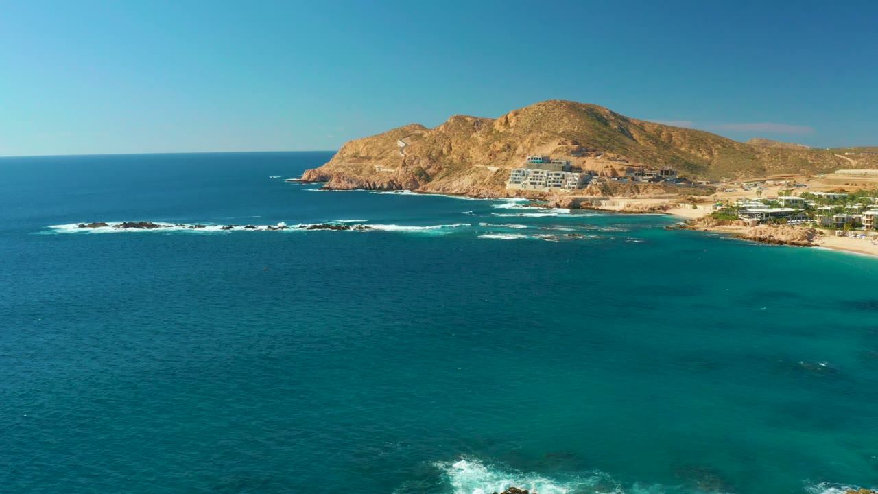 Aerial view over Chileno Beach Los Cabos with dark blue ocean at left, turquoise shallows at right, white surf along the rocks, tan hills beyond the shore, and no visible people.