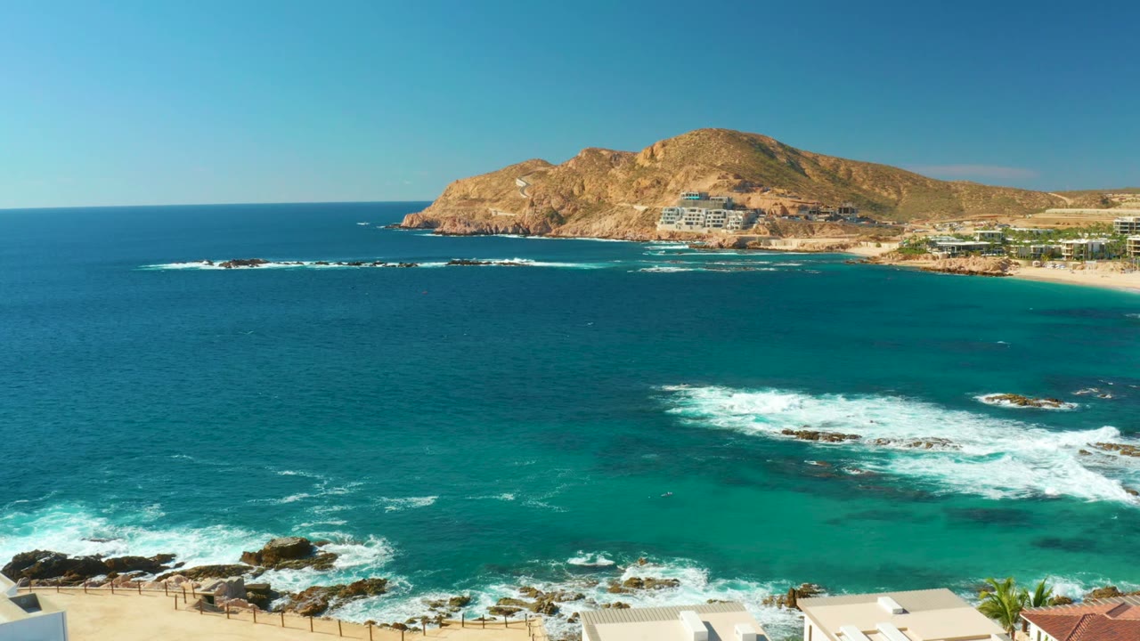 Aerial view over Chileno Beach in Los Cabos with turquoise water in the foreground, white surf on rocks, tan hillside beyond the bay, and no visible people.