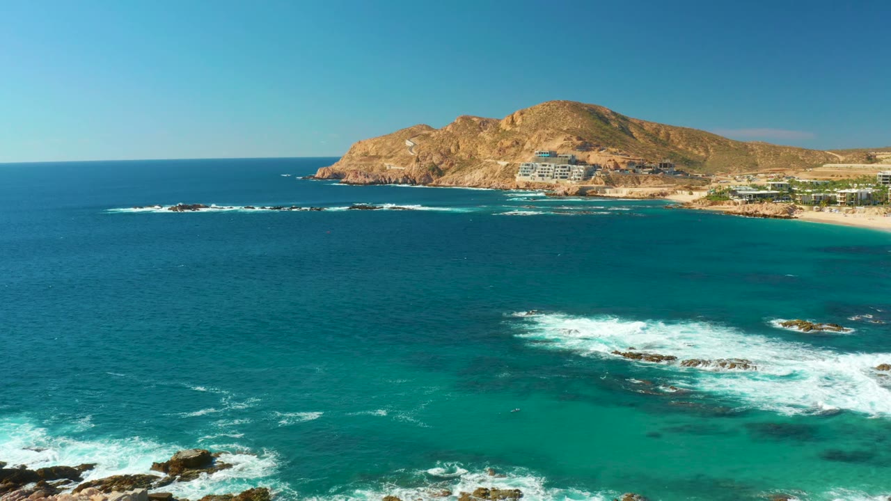 Chileno Beach shoreline from above, with deep blue water at left, turquoise shallows near the rocks, white foam spreading across the lower frame, and no visible people.