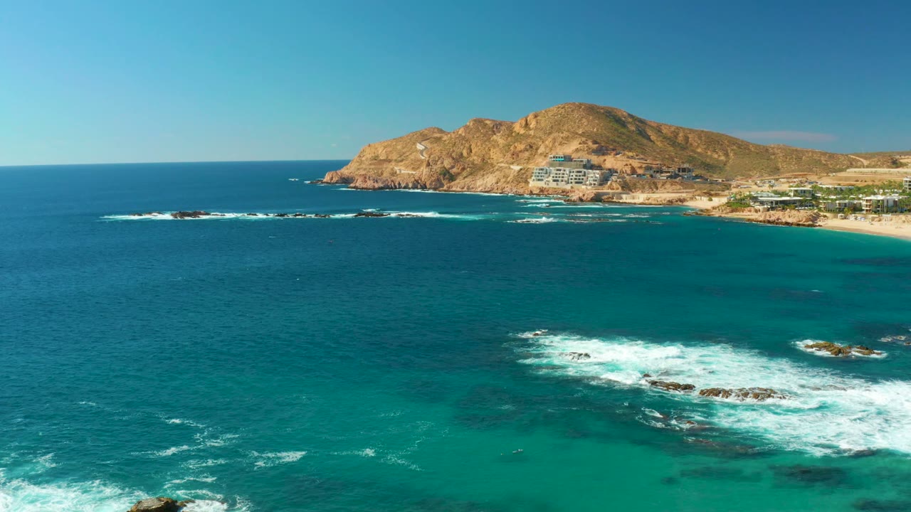 Aerial frame of Chileno Beach with a broad blue sea, breaking white waves over dark offshore rocks, a tan hill on the horizon, and no visible people.