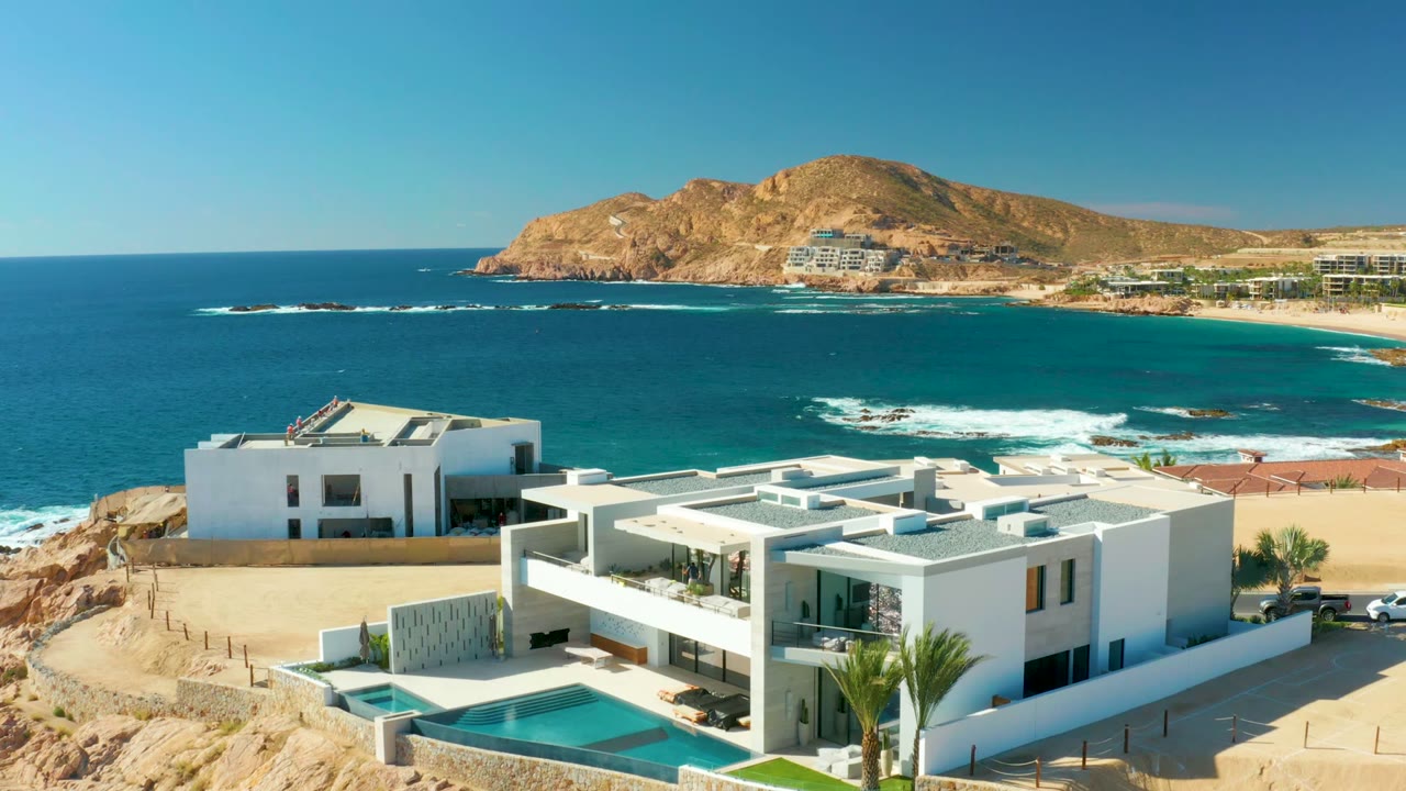 Opening aerial view at Chileno Beach Los Cabos showing white modern buildings, a pool, blue ocean, rocky surf, sandy shoreline, brown hills, and no visible people.