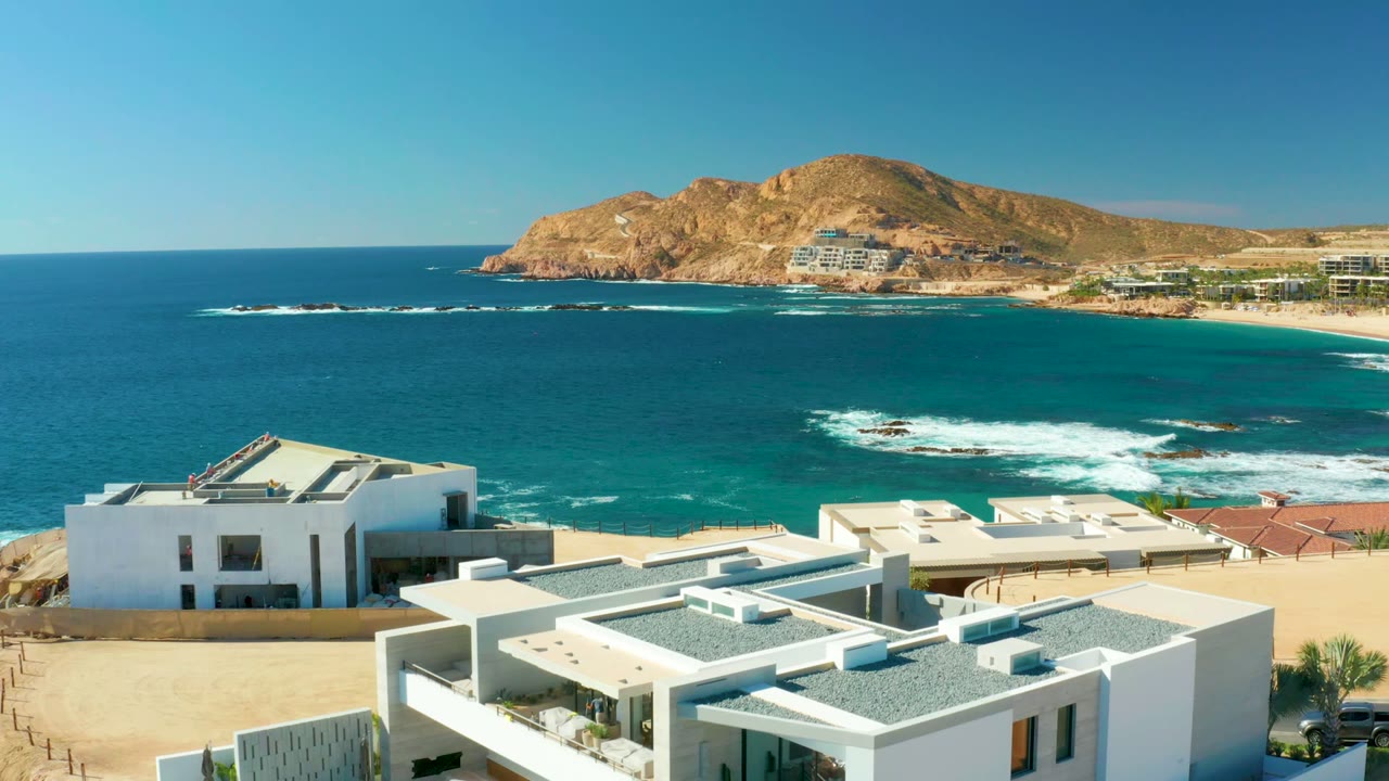 Chileno Beach Los Cabos aerial frame with white buildings lower right, a sandy beach edge, blue ocean with white surf over rocks, rocky hills across the bay, and no visible people.
