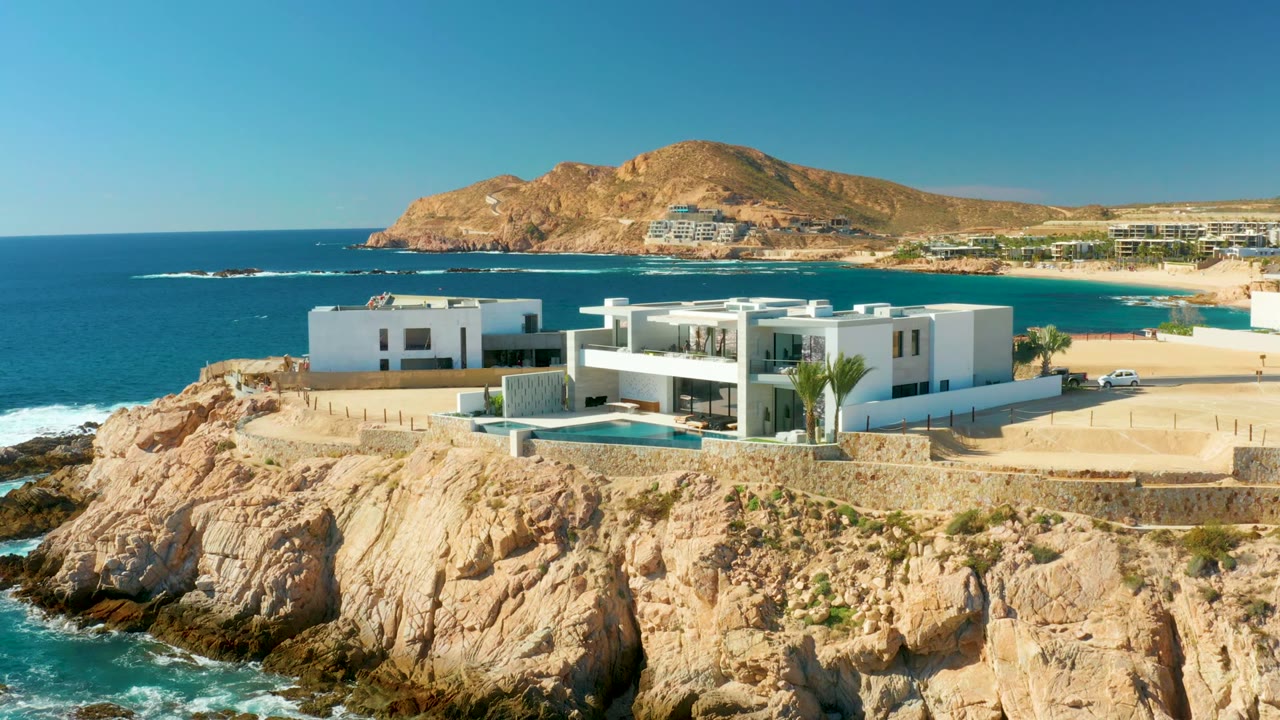 At Chileno Beach Los Cabos, a modern white villa sits above rocky cliffs and blue ocean water, with a pool and no visible people.