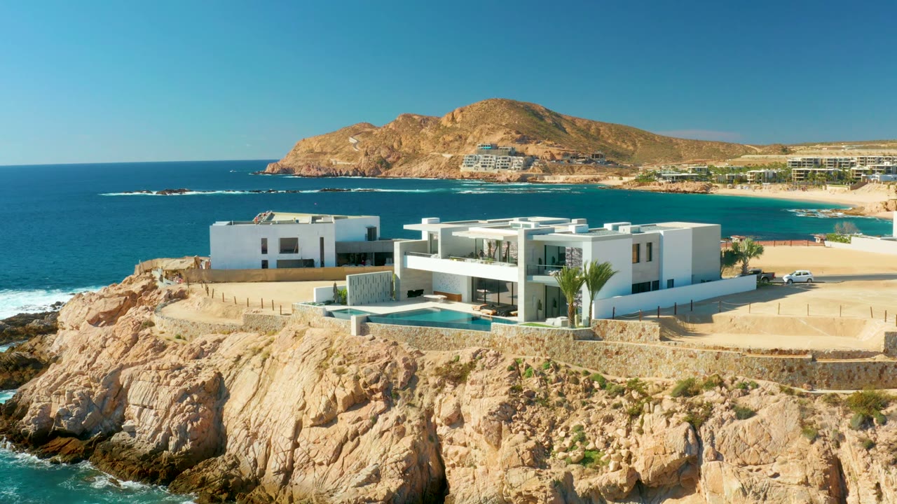 White coastal villa, pool, road, ocean, and distant hill.