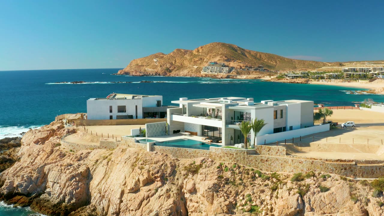 A white car is visible on the road beside the modern villa at Chileno Beach Los Cabos, with the pool, rocky cliff, beach, blue ocean, and no visible people.