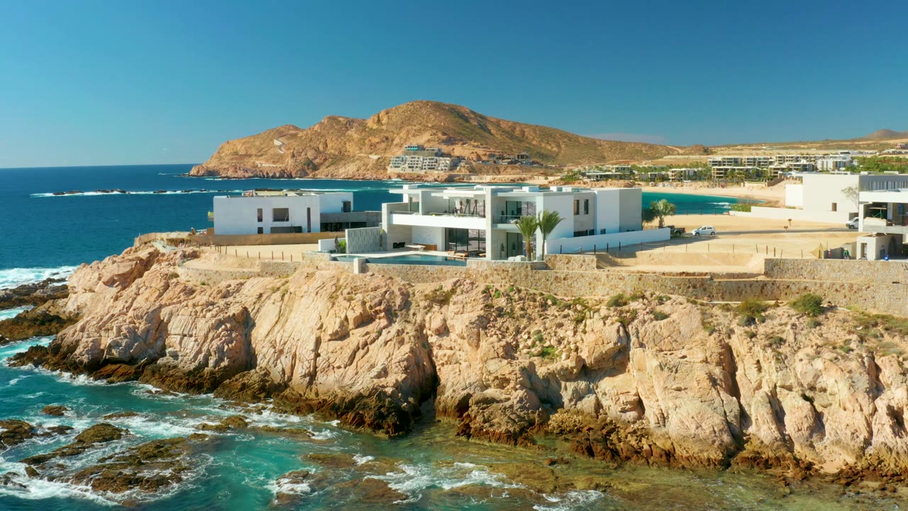 Wide aerial frame in Chileno Beach Los Cabos showing the white buildings slightly more centered over the rocky shore, with surf moving below and no visible people.