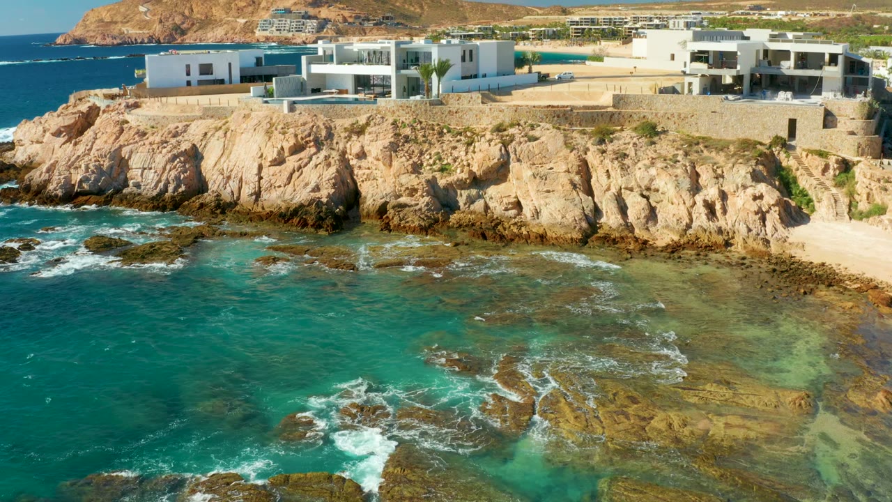 At Chileno Beach Los Cabos, white modern buildings line a rocky cliff above turquoise ocean water and reef rocks, with dry hills behind and no visible people.