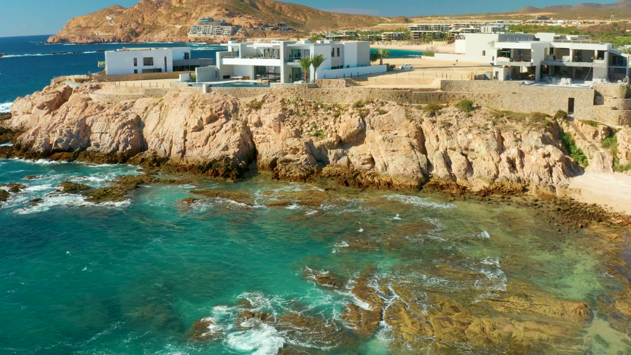 Cliffside white buildings above shallow turquoise ocean and rocks.