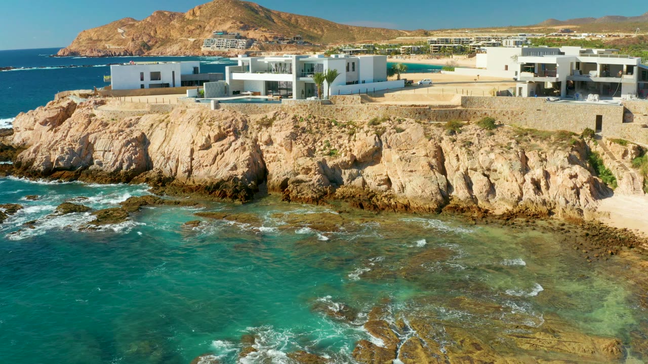 At Chileno Beach Los Cabos, the frame shows modern white buildings and a tan rock face over blue-green ocean water, with a distant hill and no visible people.