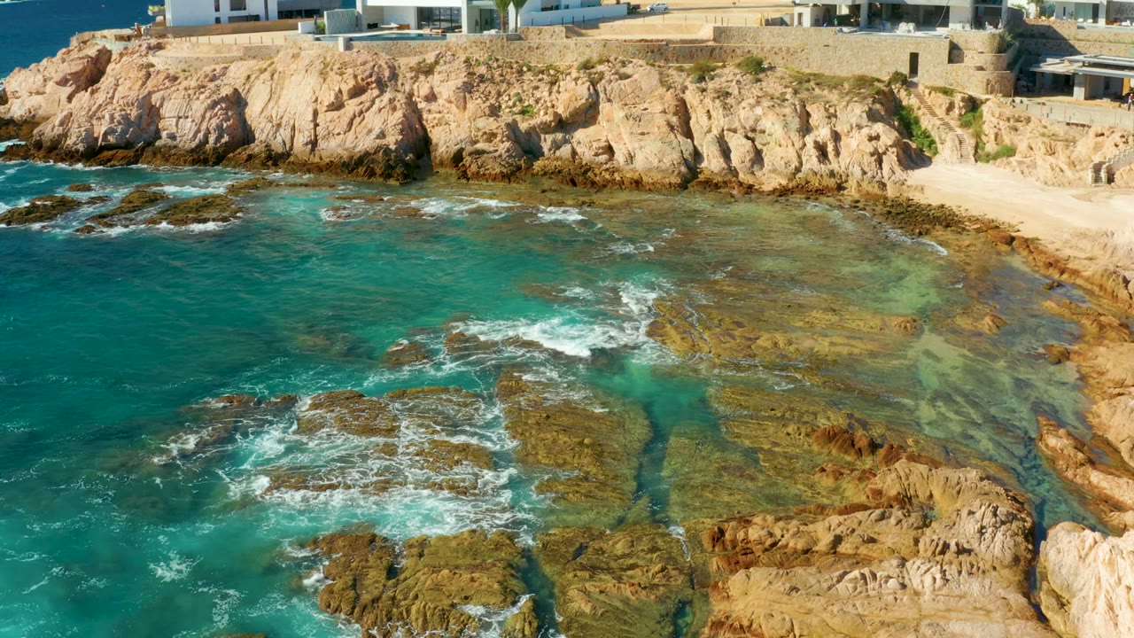 Aerial view at Chileno Beach Los Cabos showing turquoise water, flat coastal rocks, tan cliffs, and white cliffside buildings with no visible people.