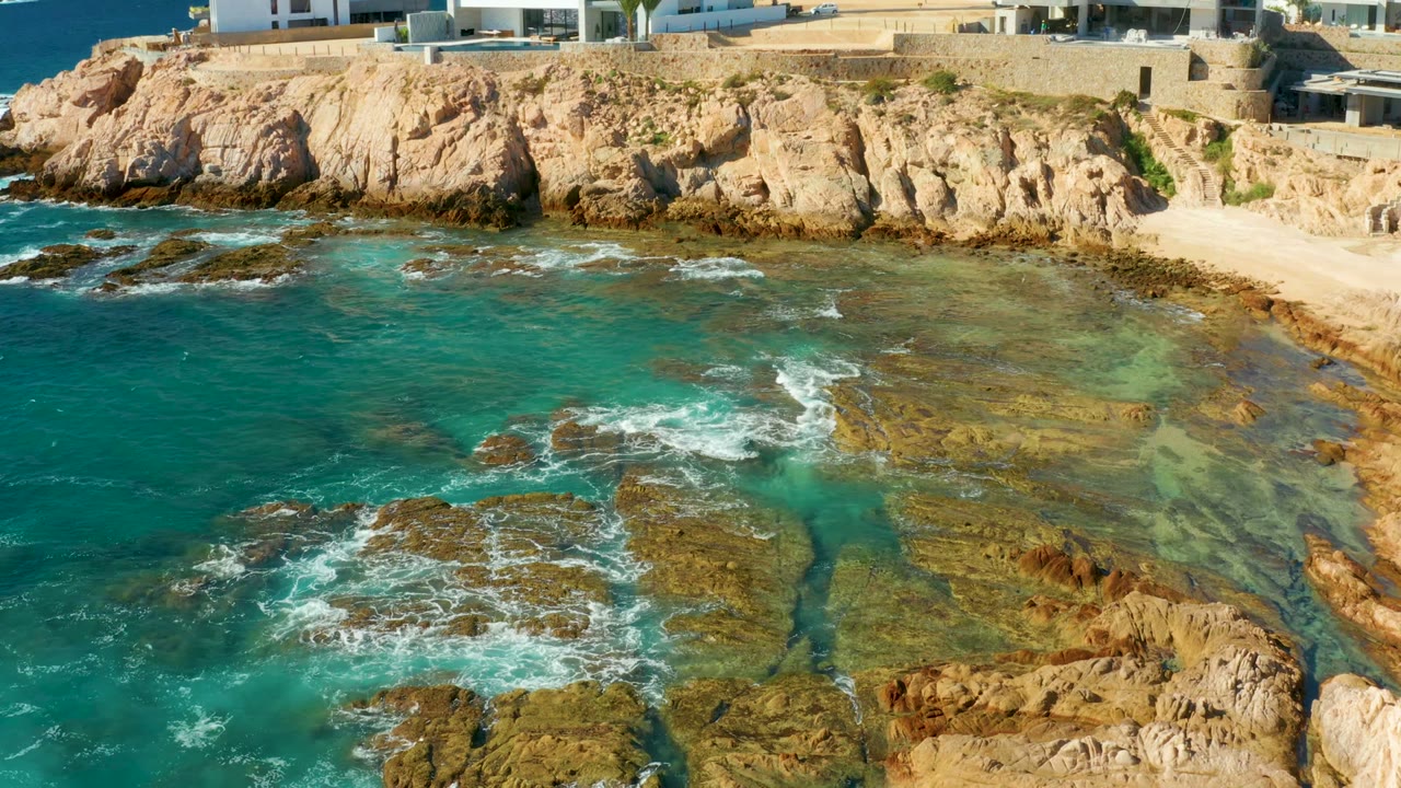 Rock shelves and white foam below tan cliffs and modern buildings.