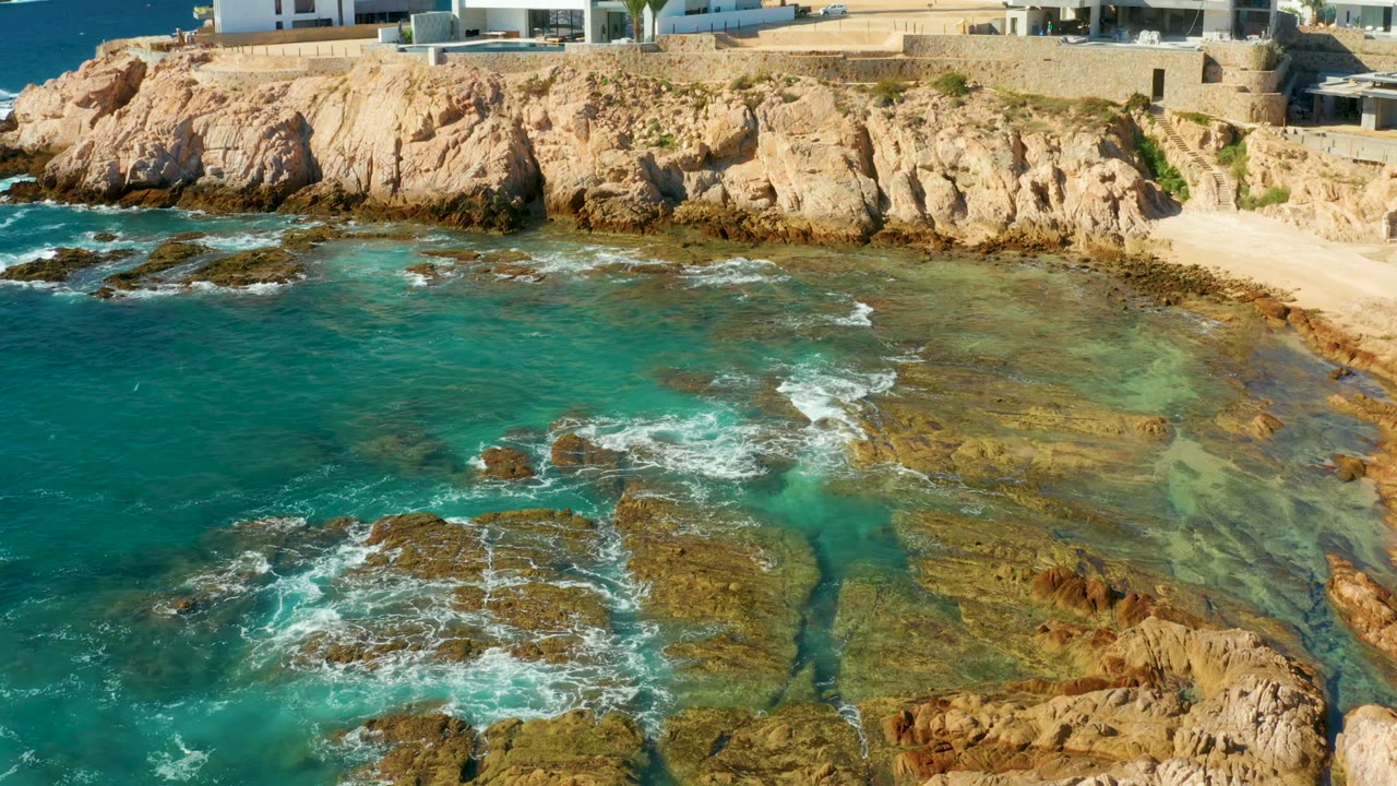Chileno Beach Los Cabos shoreline viewed from above, with clear green-blue water over reef rocks, a small sandy pocket near the cliff, and no visible people.