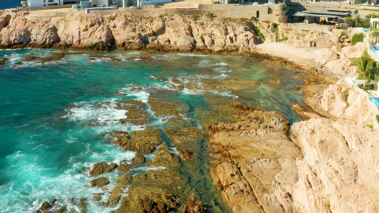 Opening view of Chileno Beach in Los Cabos with turquoise water washing over tan rock shelves, pale cliffs, a small sandy shore, distant buildings, and no visible people.
