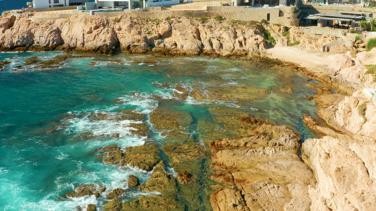 At 0:02, the aerial view holds on Chileno Beach as the largest foam patches sit left of center, with tan rock ledges in the foreground, pale cliffs and a small sandy pocket behind them, and no visible people.