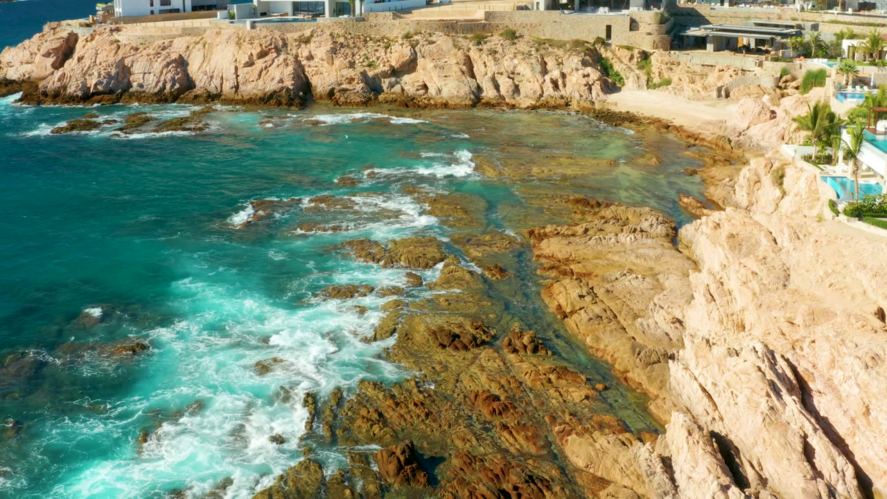 Aerial frame at Chileno Beach in Los Cabos showing turquoise surf foaming across exposed rocks below pale cliffs, with no visible people.