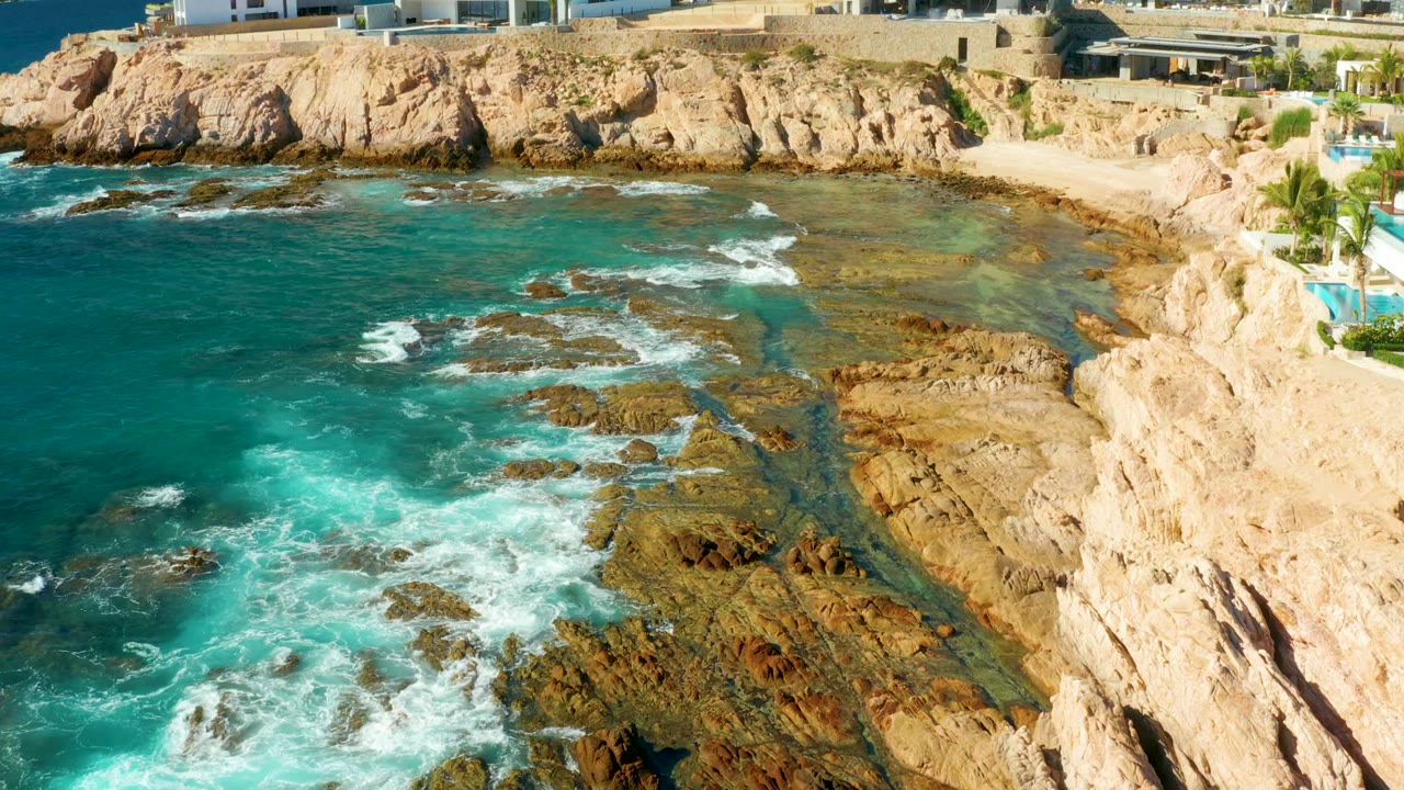 At 0:02, waves wrap around exposed offshore rocks and shallow ledges below the Chileno Beach cliffs, with no visible people in the frame.