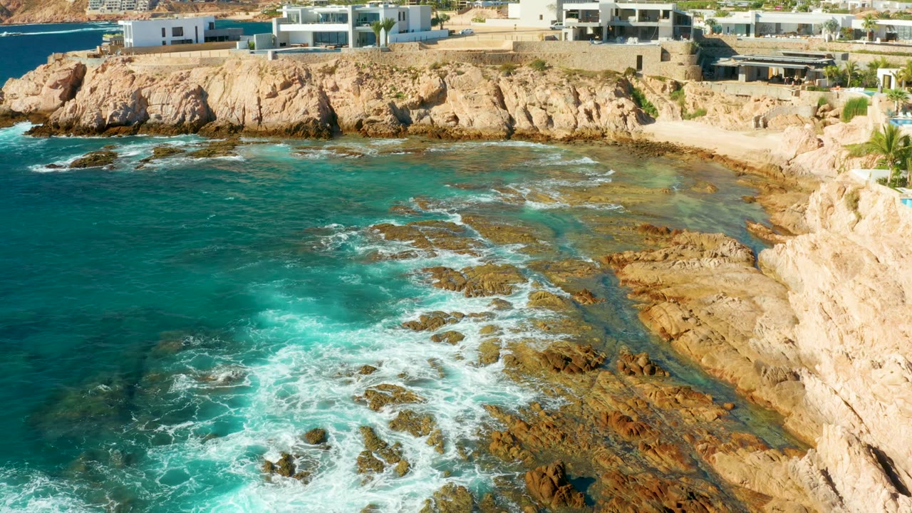 Aerial view at Chileno Beach, Los Cabos showing turquoise surf foaming across brown rock shelves beneath pale cliffs and modern buildings, with no visible people.