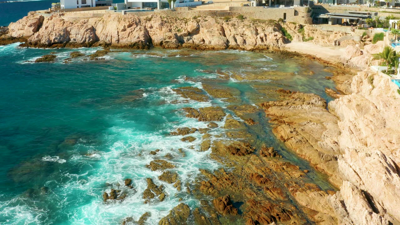Aerial view over Chileno Beach, Los Cabos with surf curling through channels in the brown rocks, clear turquoise water offshore, cliffside buildings above, and no visible people.