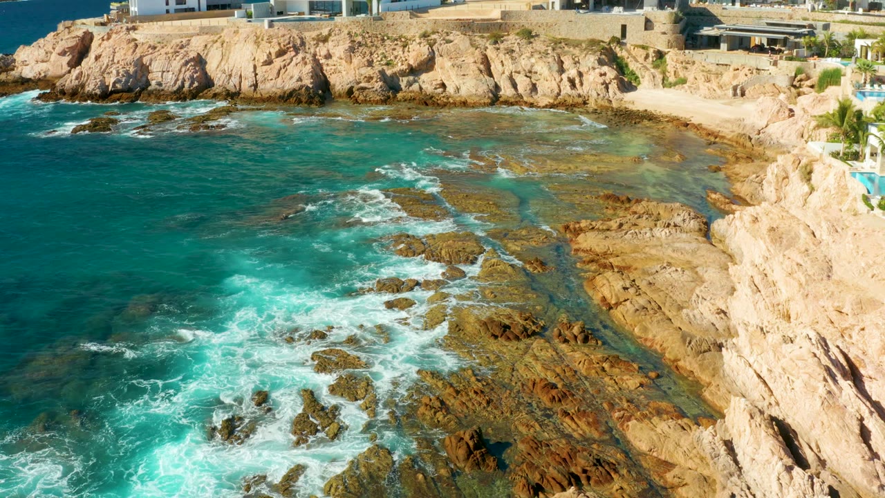 Aerial coastline with foamy waves, rocky shelf, and buildings above cliffs.