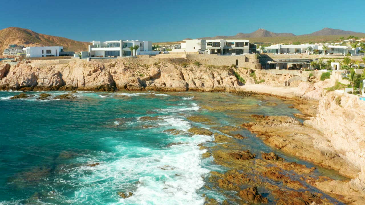 Wide elevated view of Chileno Beach in Los Cabos with turquoise water, white surf over rocks, cliffside white buildings, distant hills, and no visible people.