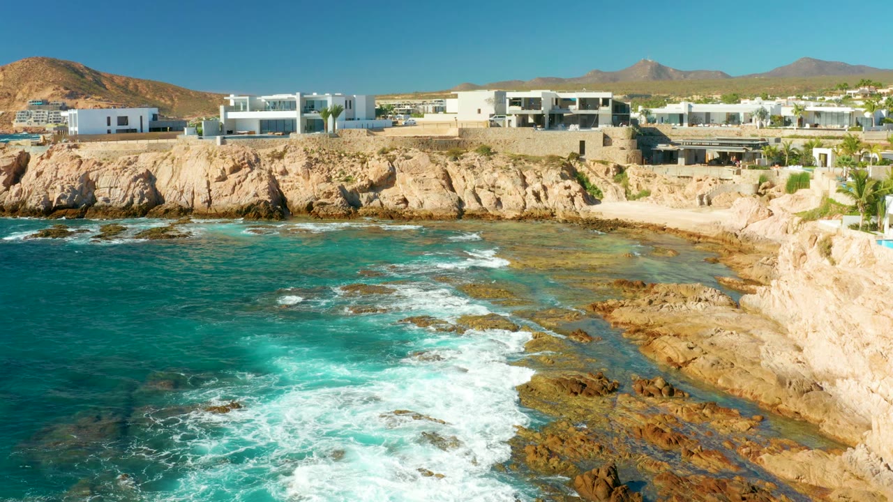 Turquoise surf breaks across rocks below white cliffside buildings.